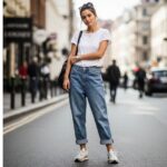 Młoda kobieta w jasnych boyfriend jeans, białej koszulce i sneakersach stoi na ulicy w miejskim otoczeniu.